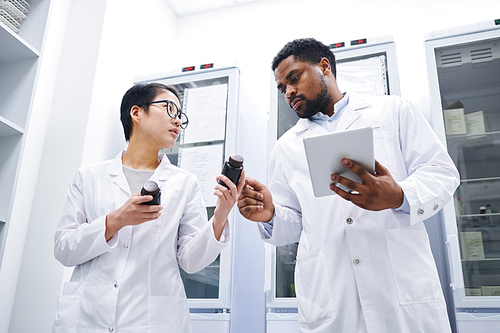 Below view of serious multi-ethnic pharmacists in white coats standing in warehouse of hospital and keeping records while viewing medications