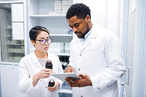 Serious thoughtful multi-ethnic pharmacists in lab coats standing in drugstore and discussing new medications while using tablet to search for information in database