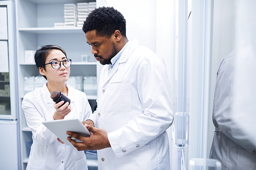 Young Asian female medical assistant in glasses showing pill bottle to black doctor and asking him about pills in storage room