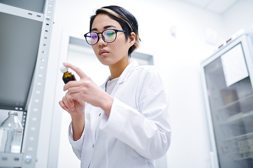 Serious concentrated young Asian pharmacist in eyeglasses wearing white coat standing in storeroom and checking expiration date on pill bottle