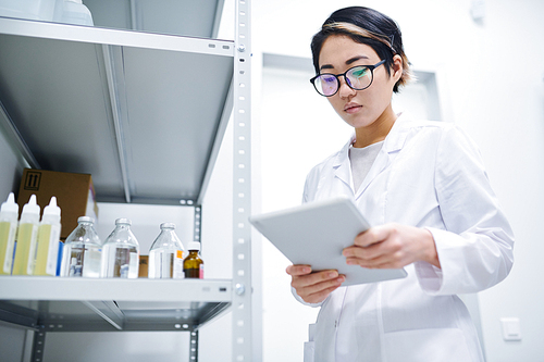 Serious concentrated young Asian lady in white coat wearing eyeglasses using digital tablet while searching for information about medication in database, she working in medical storage room