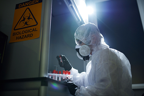 Serious concentrated microbiologist in biohazard suit and respirator examining hazardous samples while doing research in dark room