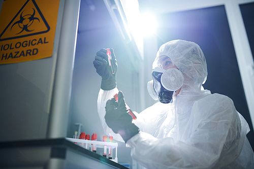 Concentrated medical scientist in biohazard suit, respirator and safety goggles sitting at desk and analyzing samples in test tube while studying dangerous virus in blood