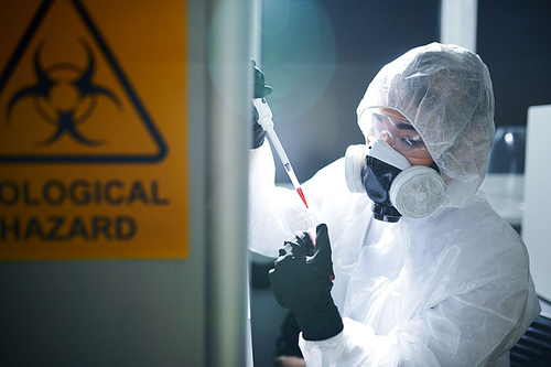 Concentrated laboratory scientist in protective workwear, rubber gloves, goggles and respirator complying with safety while working with infected substance