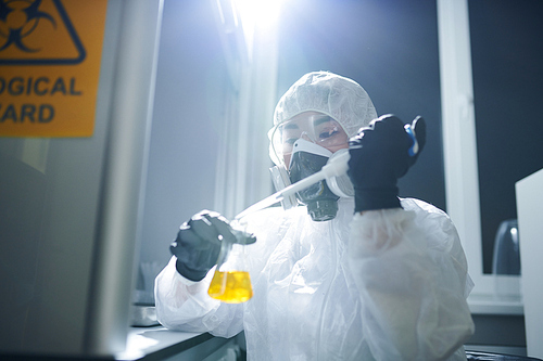 Concentrated microbiologist wearing protective goggles, biohazard suit and respiratory mask dropping reagent into flask while studying substance in laboratory