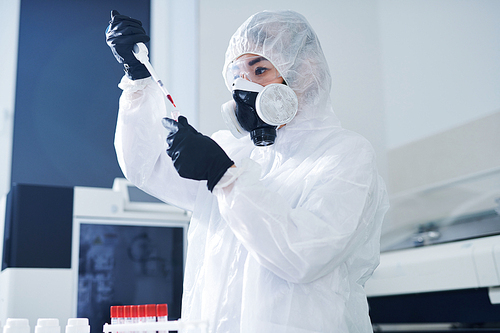 Serious concentrated healthcare scientist in biohazard suit and respirator standing in laboratory and dropping blood while testing virus sample