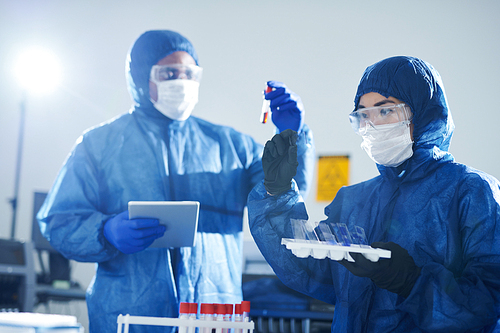 Busy virologists in blue protective suits and masks doing blood tests infected with virus: Asian lady viewing sample on glass while black man analyzing blood in test tube