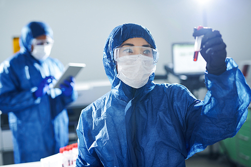 Confident healthcare scientist in blue biohazard suit and safety goggles standing in laboratory and working with dangerous sample