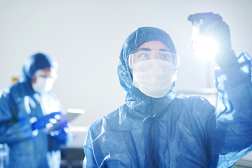 Serious pensive Asian virologist in blue biohazard suit and safety goggles standing in scientific laboratory and researching antibody in blood