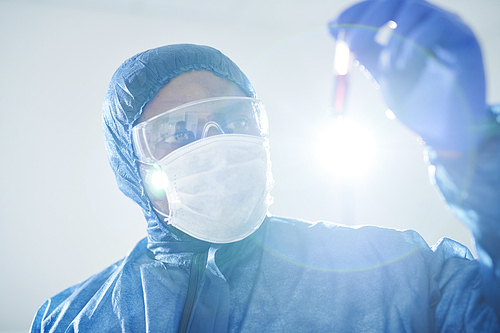 Concentrated black infectious disease scientist in biohazard suit and mask holding test tube and analyzing virus-infected blood sample