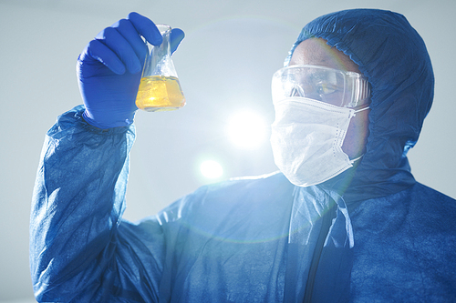 Serious concentrated black lab technician in blue protective suit and safety goggles holding flask and examining urine sample in laboratory