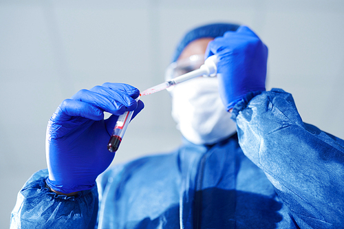 Below view of unrecognizable medical specialist in blue biohazard suit and rubber gloves doing virus research while dropping blood into test tube