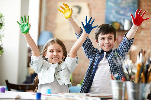 Portrait of two happy kids, boy and girl showing hands colored with paint while enjoying art class together