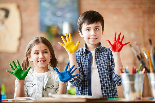 Portrait of two happy kids, boy and girl, showing hands colored with paint while enjoying art class