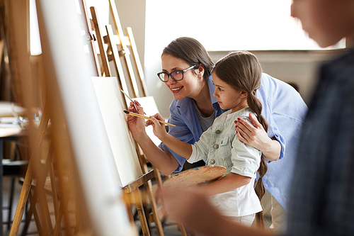 Side view portrait of smiling female teacher helping little girl painting picture on easel in art class, copy space