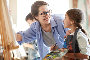 Portrait of smiling female teacher helping little girl painting picture on easel in art class, copy space