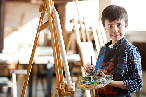 Portrait of smiling boy painting picture on easel in art class and , holding artists palette, copy space
