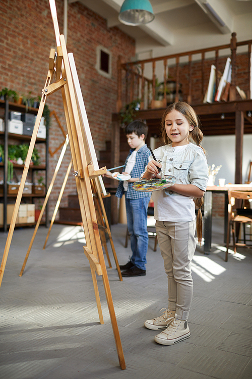 Full length portrait of cute little girl painting picture on easel in art class and holding palette, copy space