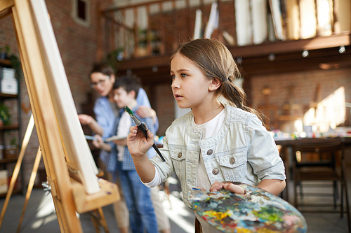 Side view portrait of focused little girl painting picture on easel in art class and holding palette, copy space