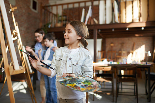 Side view portrait of joyful little girl painting picture on easel in art class and holding palette, copy space