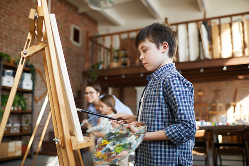 Side view portrait of smart schoolboy painting picture on easel in art class and holding palette, copy space