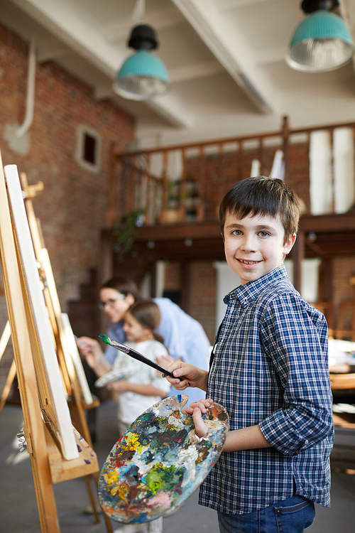 Portrait of smiling boy painting picture on easel in art class and holding palette, copy space