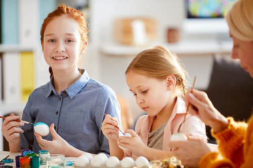 Portrait of red haired girl smiling at camera while painting eggs for Easter in sunlight, copy space