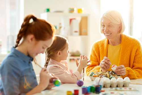 Portrait of smiling mature woman talking to little girl while painting Easter eggs together in art class, copy space