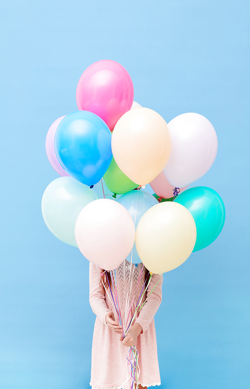 Front view portrait of cute little girl hiding behind balloons standing against pastel blue background, party concept, copy space