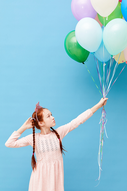 Portrait of cute red haired girl holding balloons standing against pastel blue background, Birthday party concept, copy space