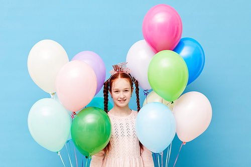 Waist up portrait of cute red-haired girl holding balloons standing against pastel blue background, Birthday party concept, copy space