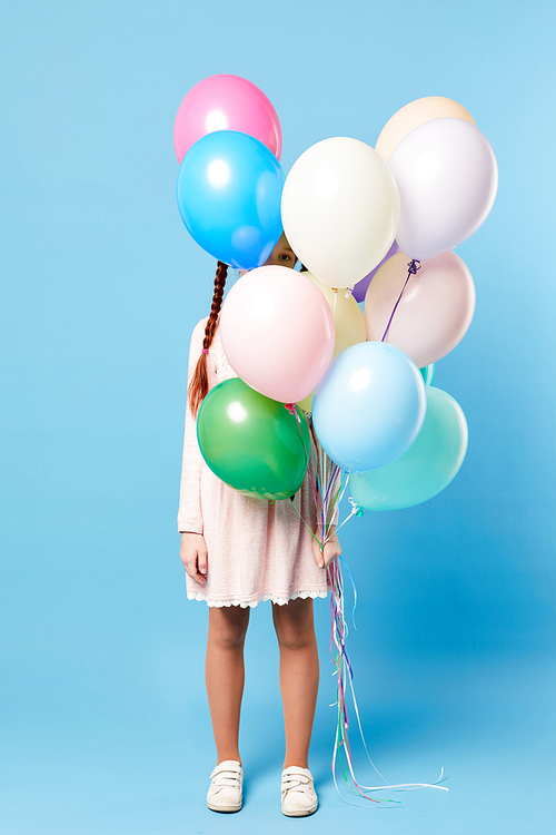Full length portrait of cute girl hiding behind balloons standing against pastel blue background, party concept, copy space