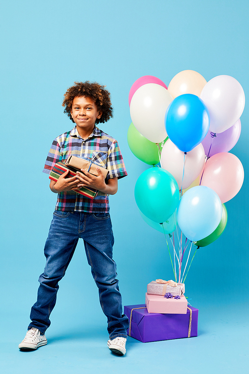 Full length portrait of cheerful African-American boy holding presents posing against blue background, Birthday party concept
