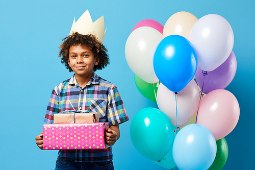 Waist up portrait of cheerful African-American boy holding presents posing against blue background, Birthday party concept, copy space