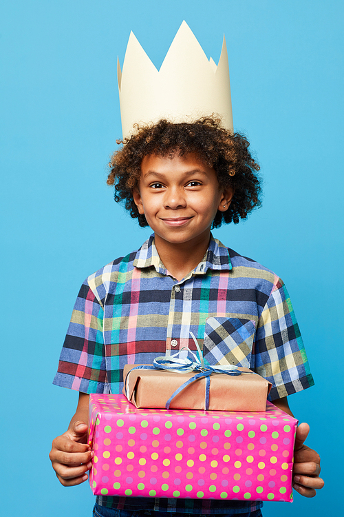 Waist up portrait of cheerful African-American boy holding presents posing against blue background, Birthday party concept