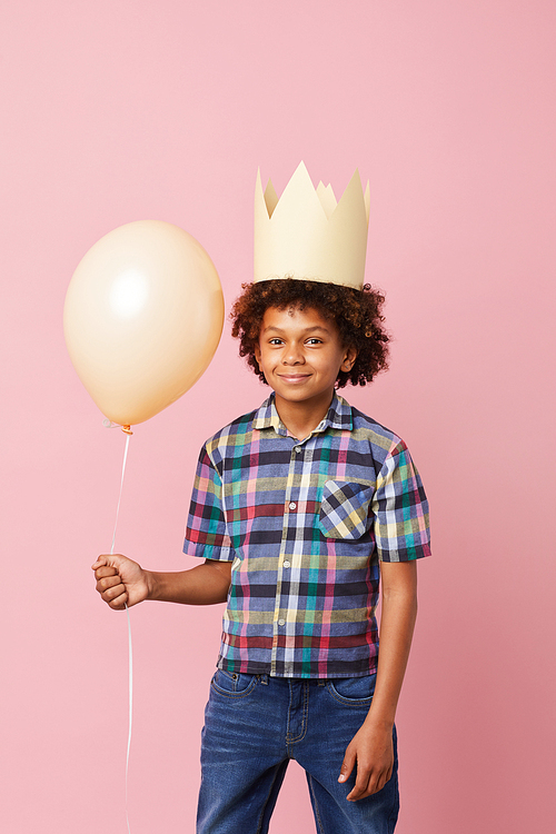 Portrait of smiling African-American boy wearing crown posing against pink background with balloon, Birthday party concept