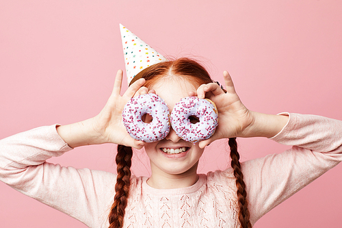 Waist up portrait of smiling red haired girl holding donuts posing against pink background, Birthday party concept