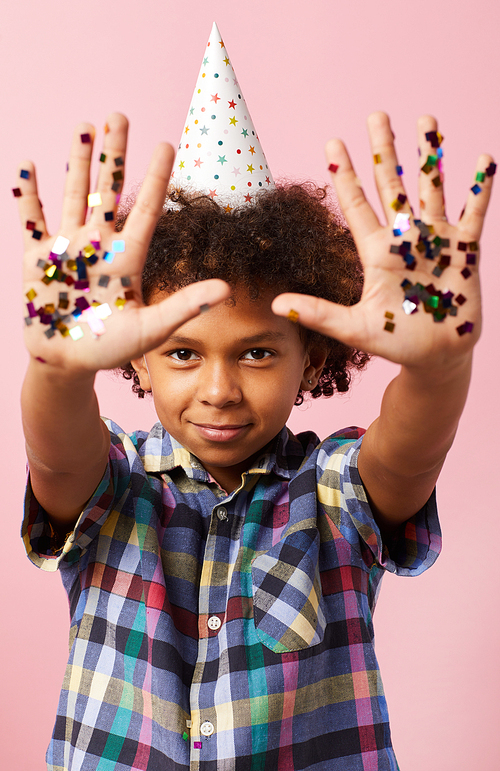 Waist up portrait of cute African-American boy showing hands with confetti at camera while posing against pink background