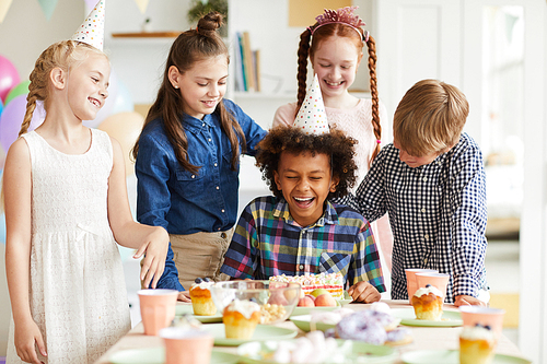 Multi-ethnic group of children celebrating Birthday standing at table together with African-American boy pulling him face in cake