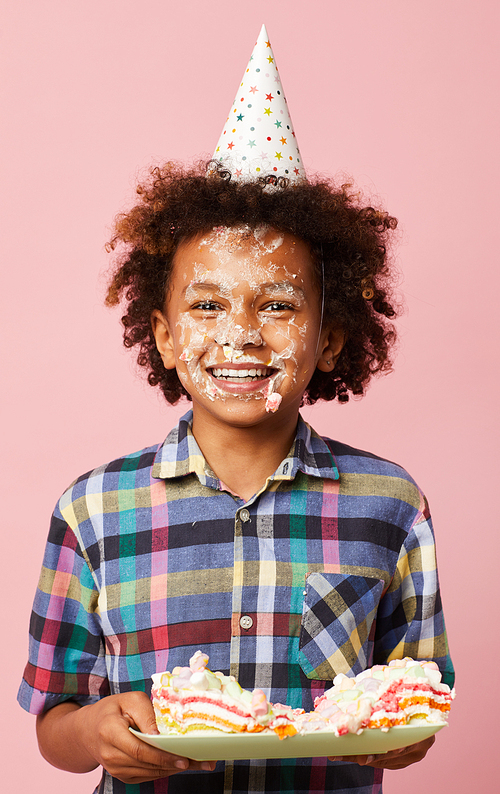 Waist up portrait of cute African-American boy holding cake and smiling at camera while posing against pink background