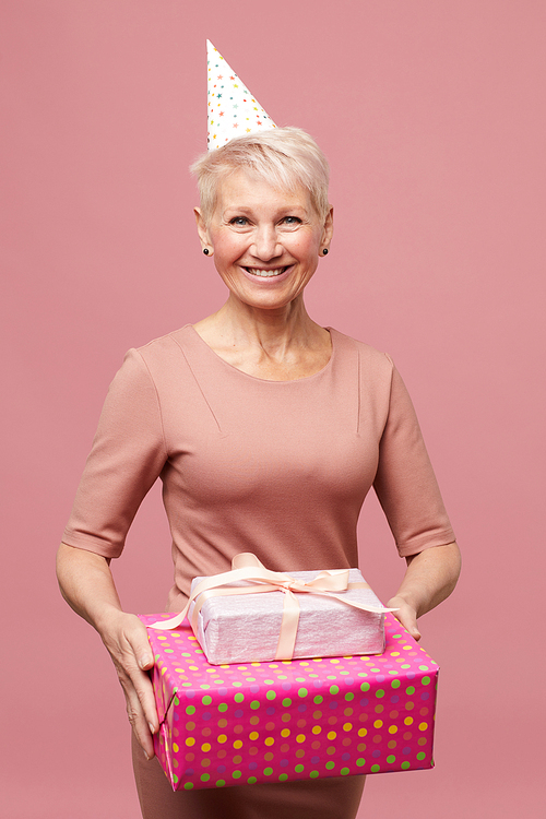 Portrait of happy mature lady with short hair wearing party hat and pink dress holding stack of birthday gifts