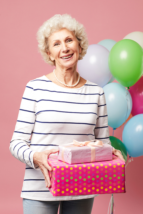 Cheerful optimistic curly-haired mature lady with necklace wearing stripped shirt standing against heap of balloons and holding gifts while enjoying birthday party