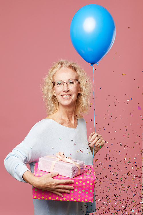 Portrait of cheerful attractive mature woman with curly hair standing against pink background and holding balloon and gifts