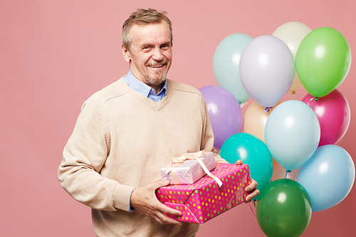 Jolly handsome mature bearded man in sweater standing against pink background with multi-colored balloons and holding birthday gifts