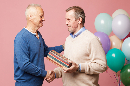 Content handsome mature man in sweater holding stripped gift box and shaking hand of friend while accepting birthday congratulations at party