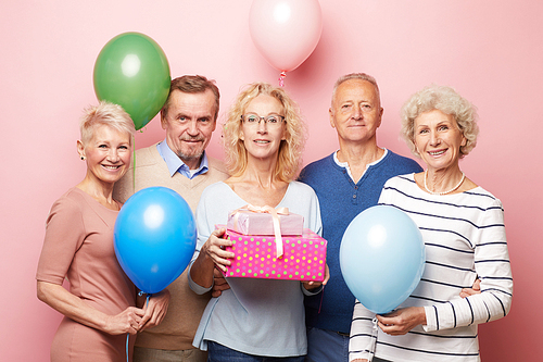 Smiling mature lady and her friends holding colorful balloons standing against pink wall and  while posing for photo at birthday party
