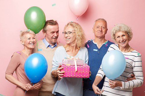 Group of positive excited mature friends in casual outfits congratulating curly-haired lady with birthday presents in hands during party