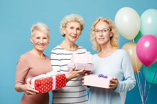 Portrait of smiling beautiful mature ladies standing against heap of balloons and blue wall and holding beautiful gift boxes, womens day concept