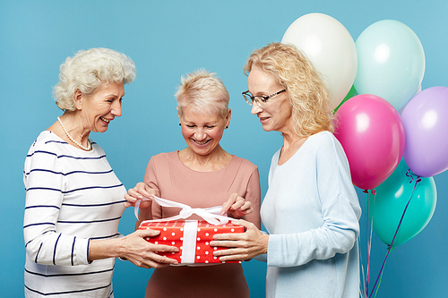 Group of positive senior ladies in casual clothing standing in studio decorated with colorful balloons and opening gift box together