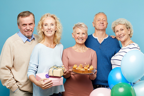 Cheerful mature friends gathering for birthday party of lady holding sweet cake, they posing together against blue background